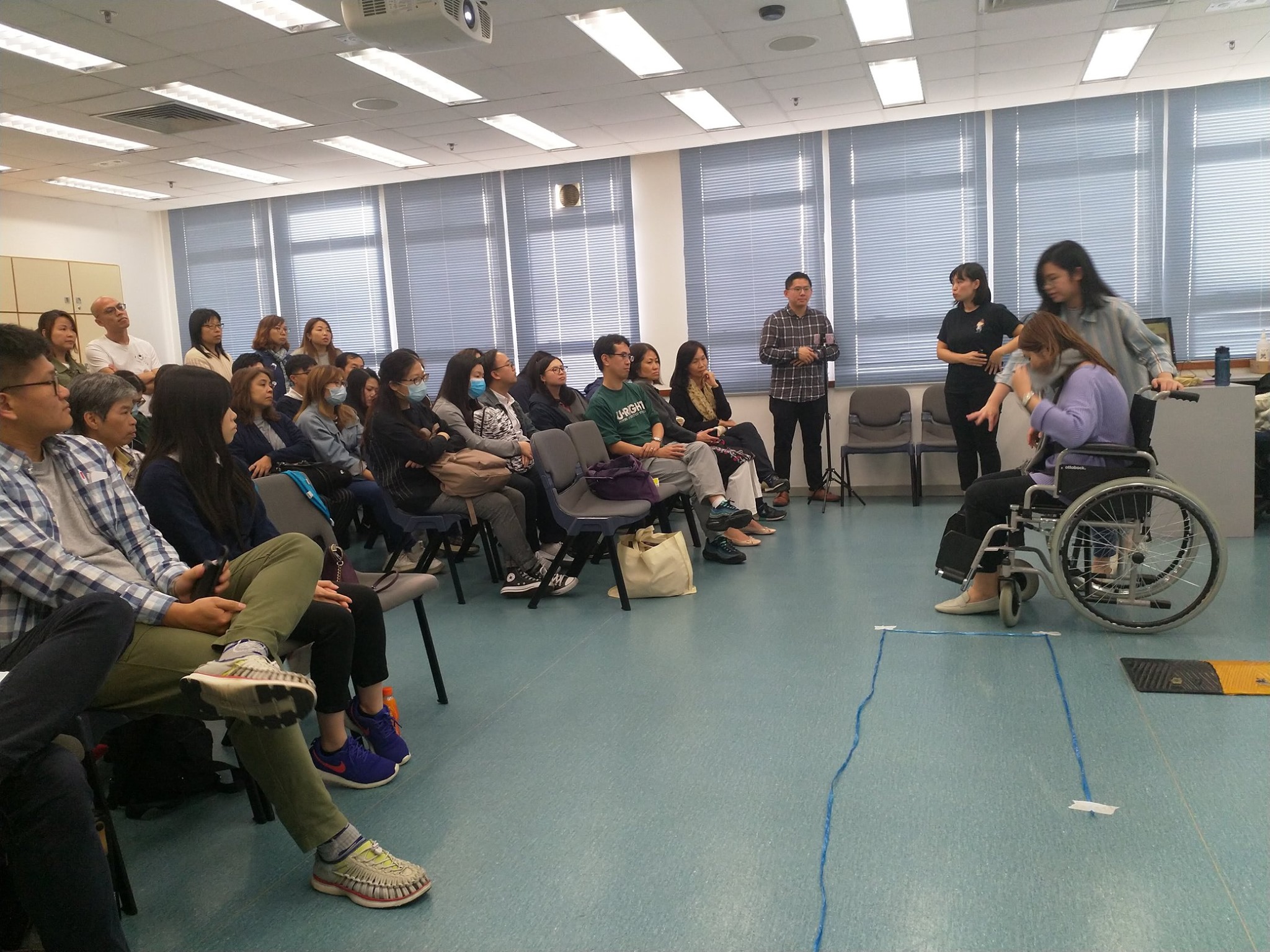 群人在教室内参加无障碍培训或讲座，听众认真观看。一名坐在轮椅上的女性正在由另一位女性协助，通过模拟无障碍通道进行演练，现场有标示线。